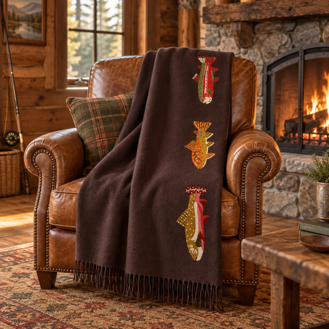 A dark brown throw blanket with embroidered fish motifs draped over a brown leather armchair in a cozy rustic living room with a fireplace.
