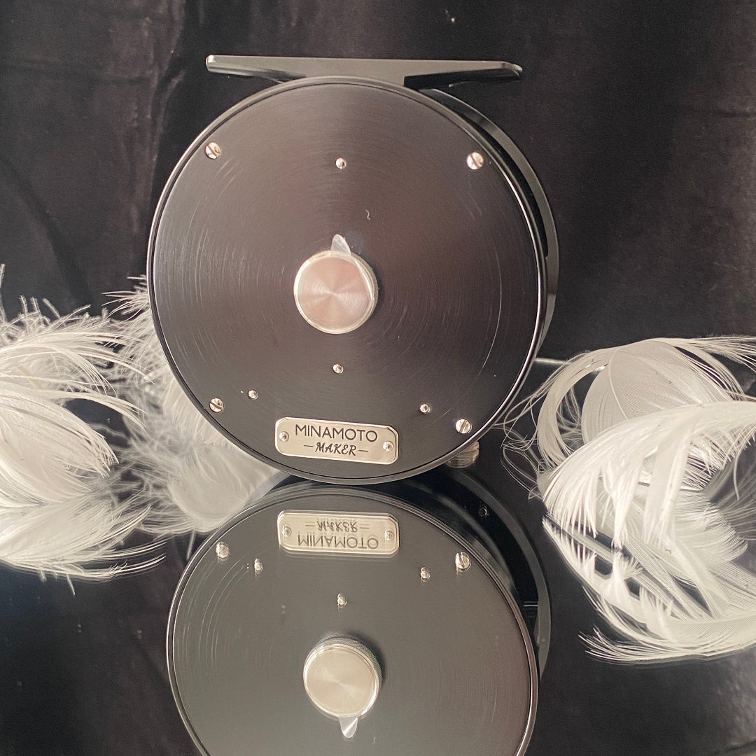 A black Minamoto Maker fly fishing reel shown from the back, featuring a brushed metal plate, silver hardware, and a silver brand plaque. White feathers are arranged on both sides of the reel against a black fabric background.