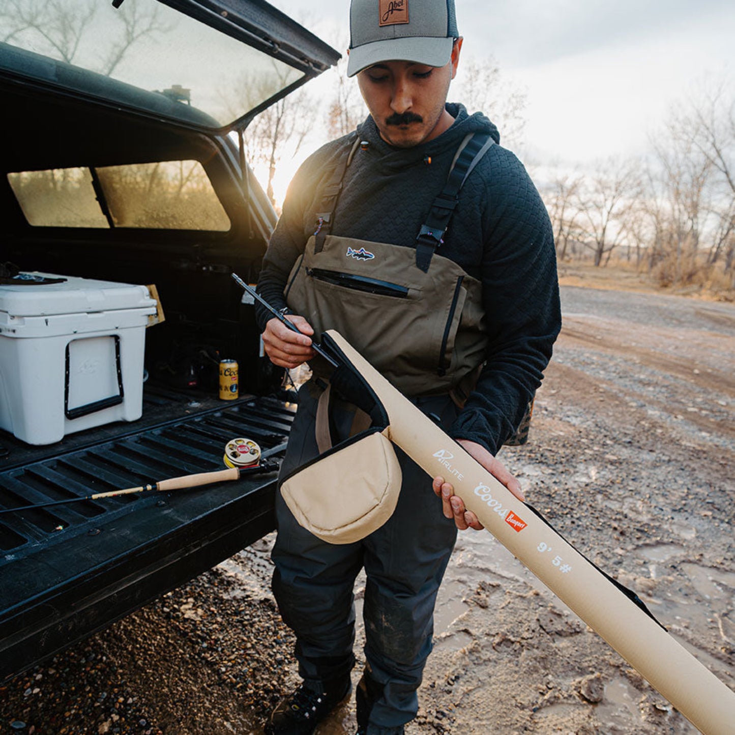 Man in hunting gear holding a bow near an open truck bed with hunting equipment.
