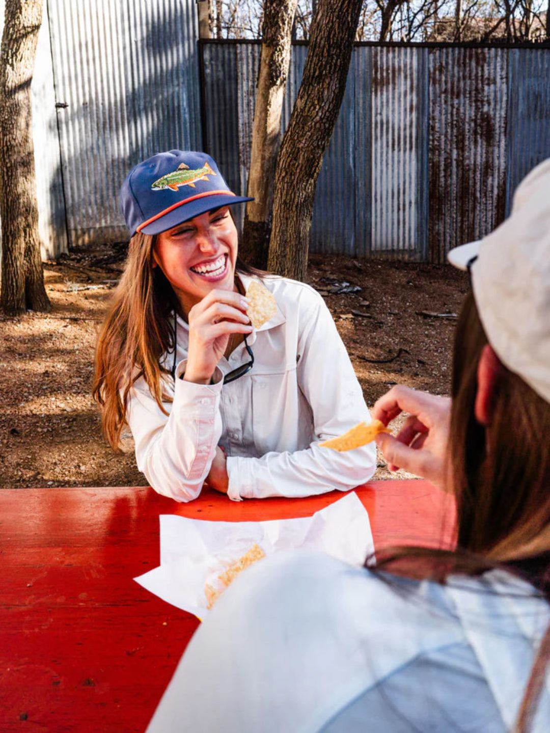A woman wearing a blue cap with a fish logo and a white long-sleeve shirt sits at a red outdoor table, smiling while eating. Another person is partially visible in the foreground.