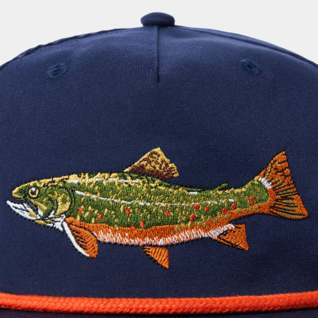 A close-up of a navy blue flat-brim cap featuring a large embroidered trout on the front and a bright orange rope detail along the brim.