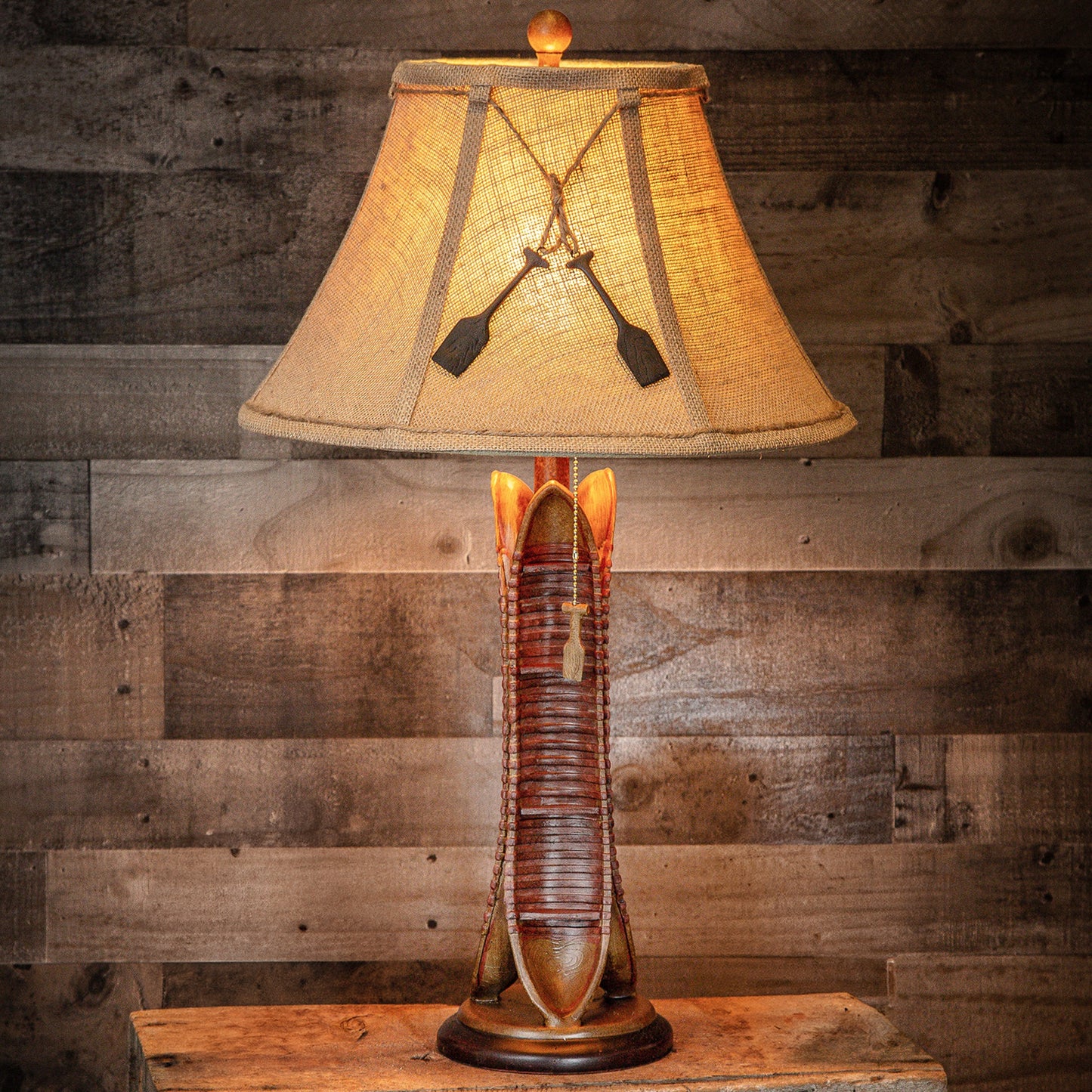 A rustic table lamp with a woven beige shade decorated with two black oars and a wooden base shaped like a boot, set against a dark wood plank wall.