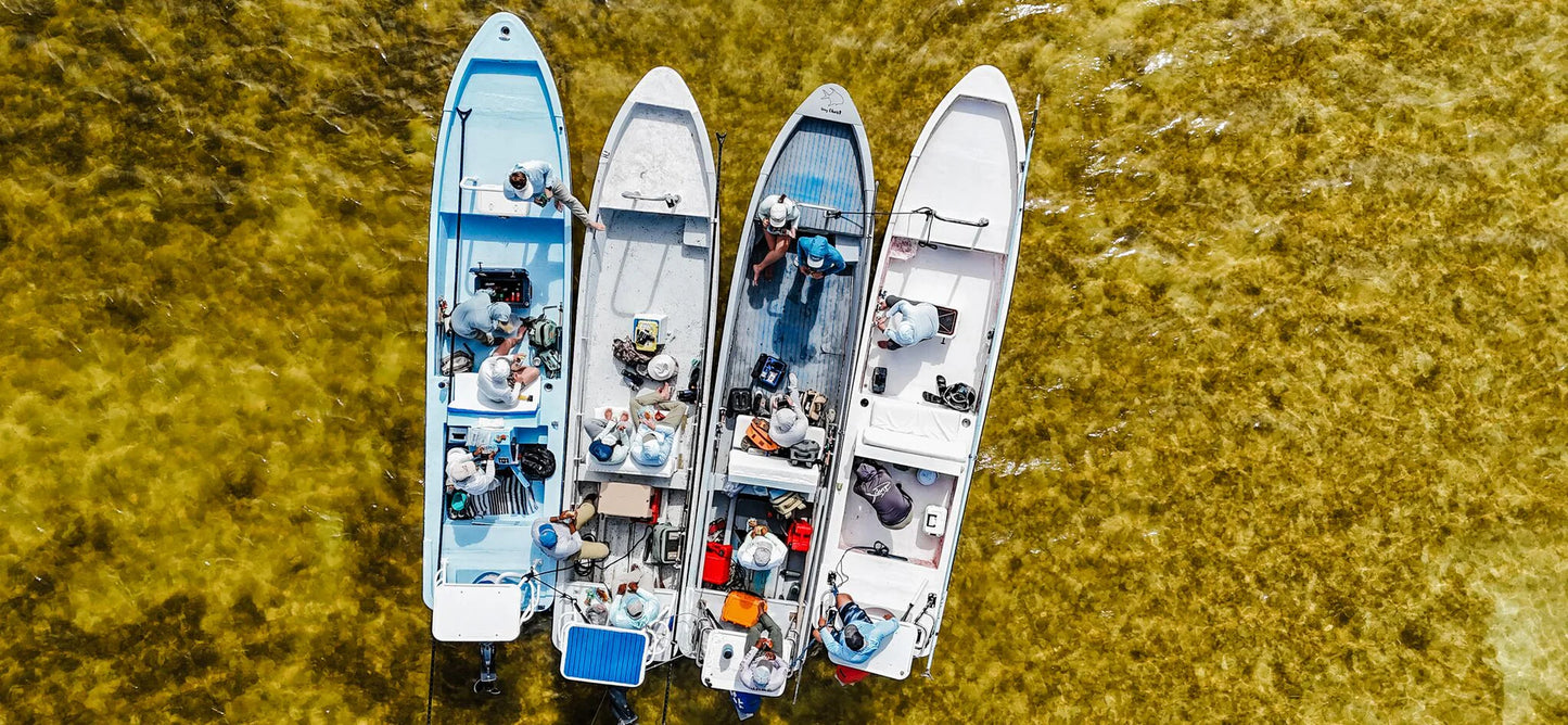 An aerial top-down view of four small white boats anchored in shallow, sunlit water with a yellow-green seabed visible beneath the surface. Several people are seated inside the boats.