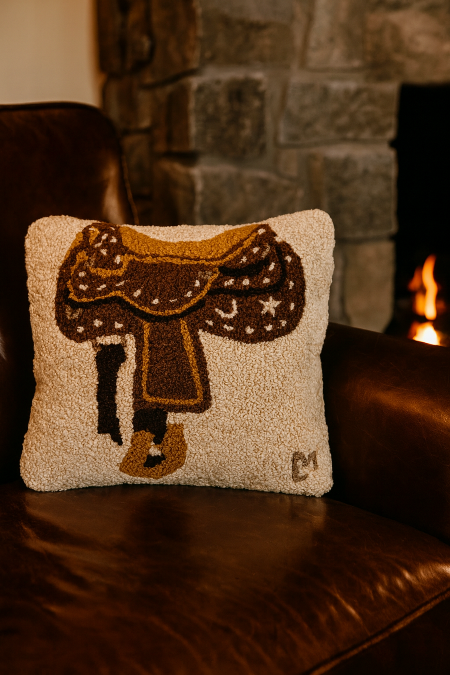 Decorative pillow with a saddle design on a leather couch in front of a stone fireplace.
