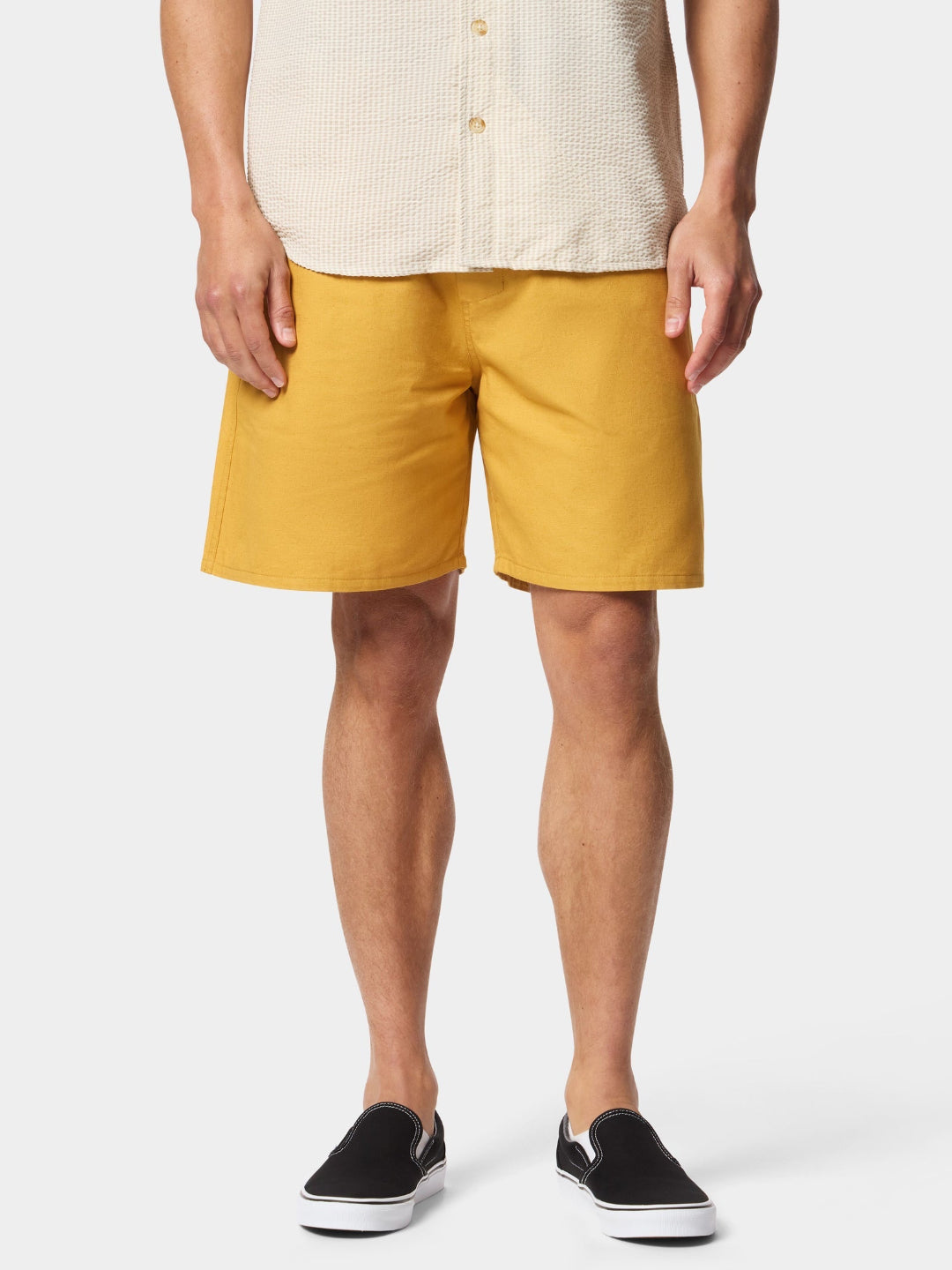 A man wearing mustard yellow shorts, a cream-colored textured short-sleeve button-up shirt, and black slip-on sneakers, shown from the waist down against a light gray studio background.