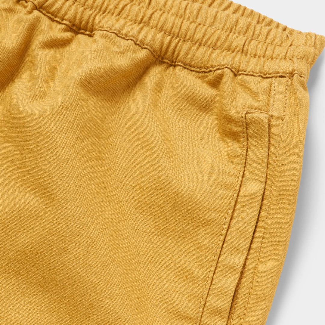 Close-up of mustard yellow fabric with an elastic waistband and a side pocket seam, photographed on a light gray background.