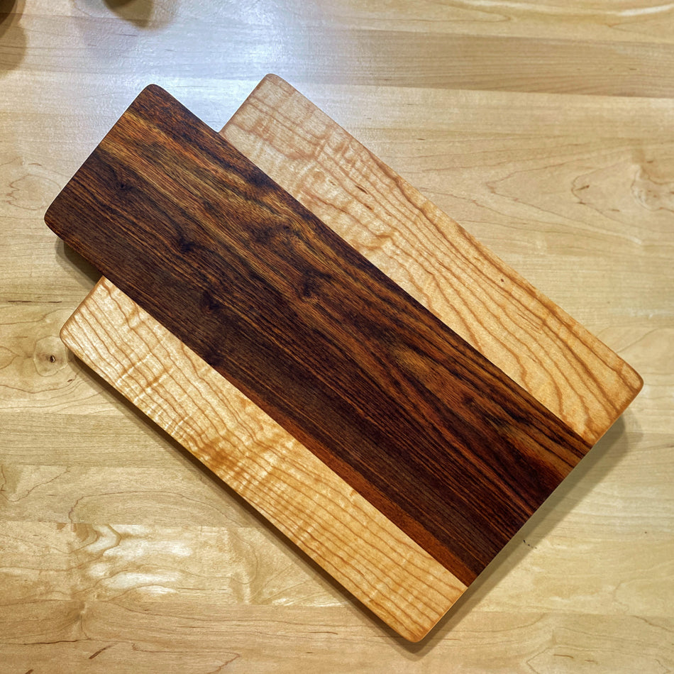 This remarkable Charcuterie Board is handmade with Caribbean Rosewood and Curly Birchwood. These exquisite woods create a stunning color combination, perfect for patio evenings with cocktails and sushi. This board is double-sided and offers an ideal size for crafting delightful charcuterie spreads.