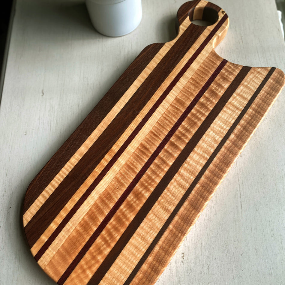 
                  
                    This handcrafted Charcuterie Board is created with Curly Birch, Walnut and Purple Heart with a Millie's All-Purpose Penetrating Tung Oil finish. It measures 17" (including its handle), 7 3/4" wide, and 1" thick, making it ideal for intimate social gatherings.
                  
                