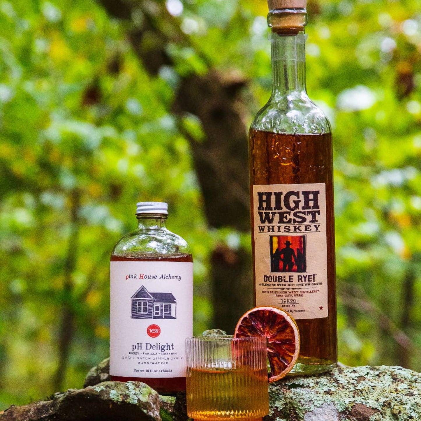 Two bottles of High West Whiskey and a glass with a drink on a rock with a forest background