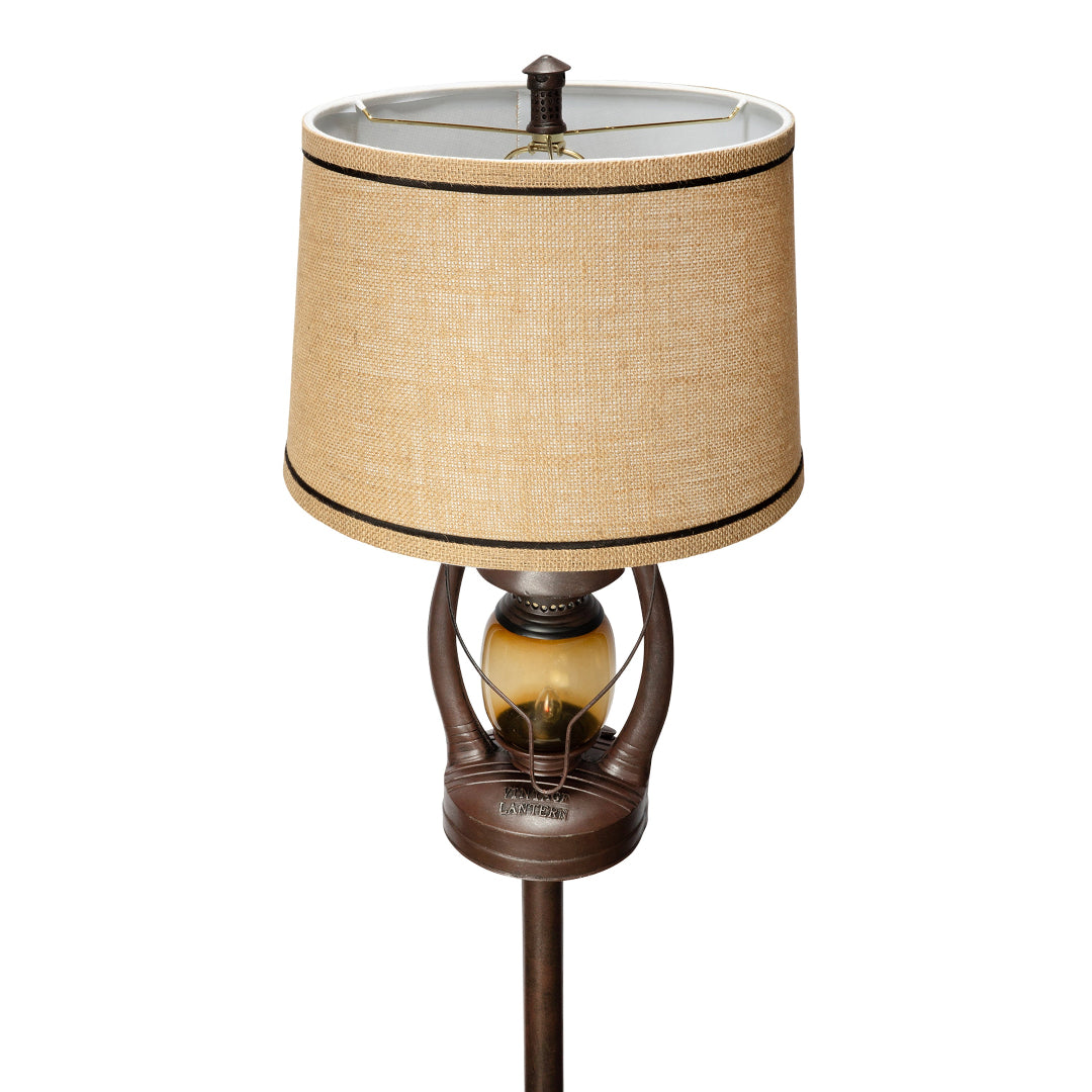 A table lamp with a textured beige fabric drum shade, a dark bronze metal base, and an amber glass lantern-style bulb holder, shown on a plain white background.