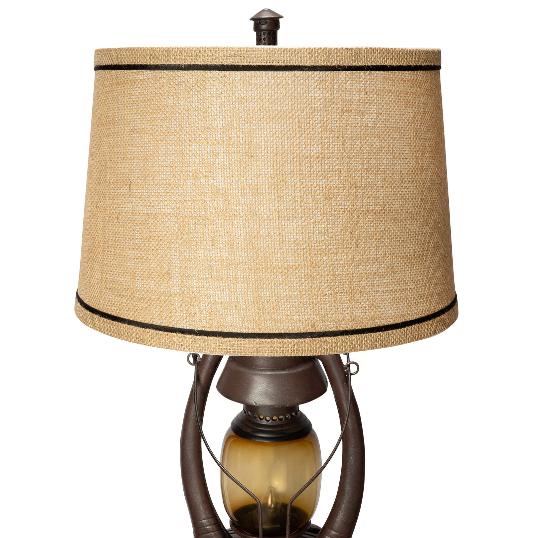 A rustic table lamp with a large beige burlap-textured drum shade trimmed with black bands, mounted on a dark brown metal frame with a glass oil lamp bulb.