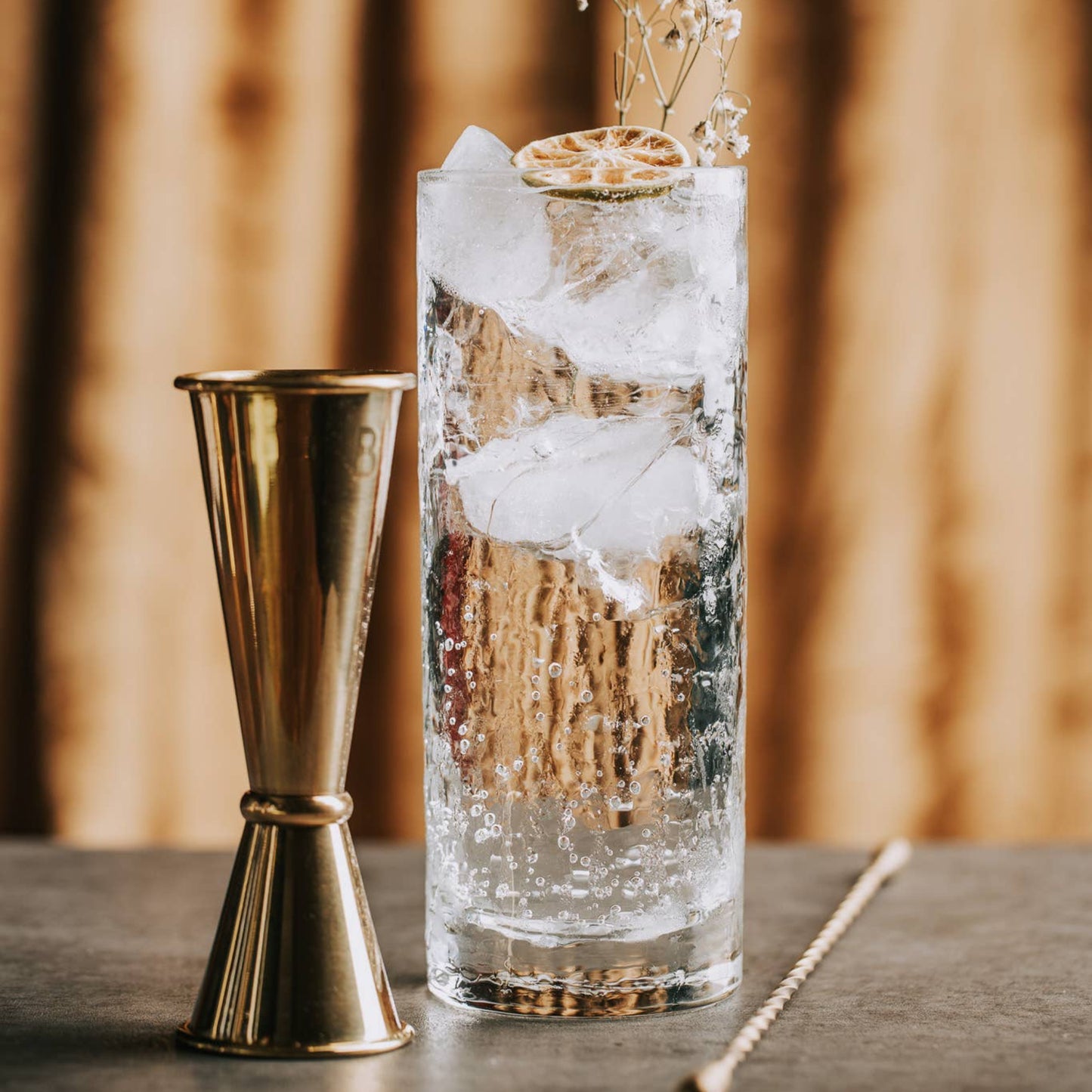 Cocktail with ice and a decorative straw on a wooden surface, accompanied by a gold jigger.