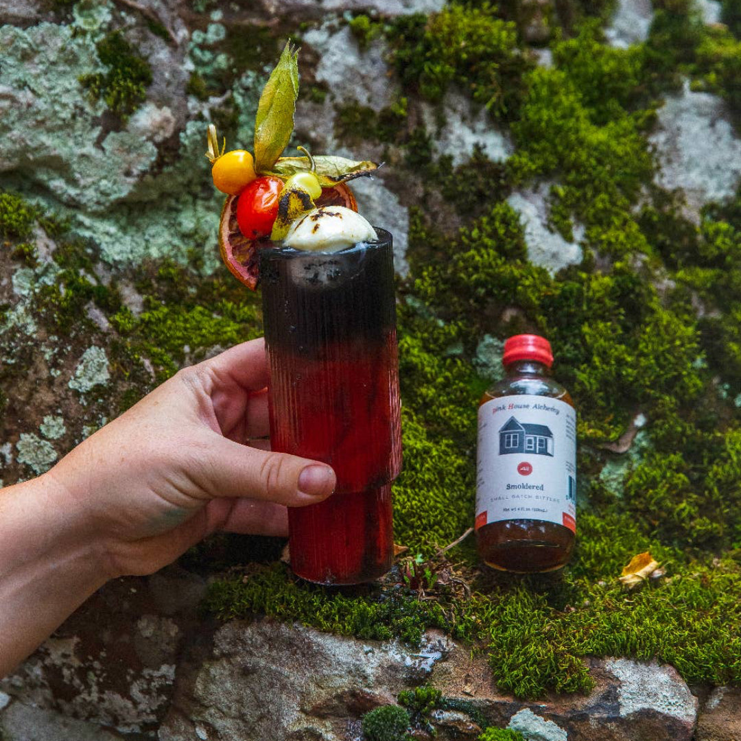 Hand holding a cocktail with garnishes next to a bottle on a mossy rock background