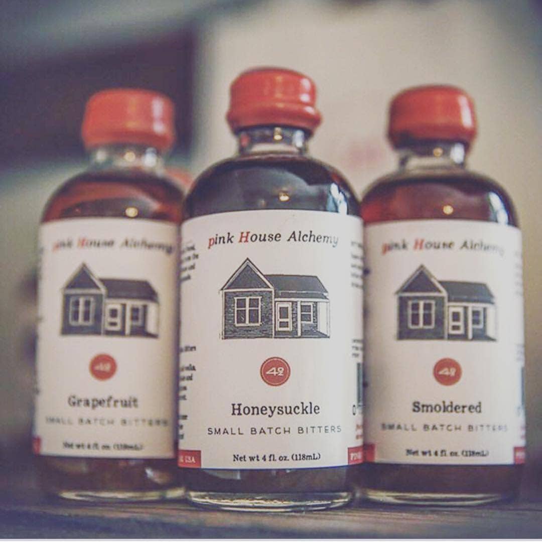 Three bottles of Pink House Alchemy bitters with red caps on a blurred background.